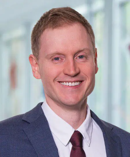 A smiling man in a suit and tie with a blurred indoor background.