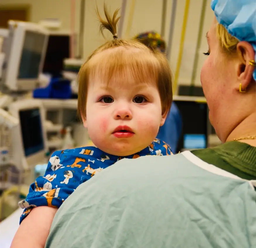 A toddler with a ponytail sits on an adult's shoulder in a medical setting.
