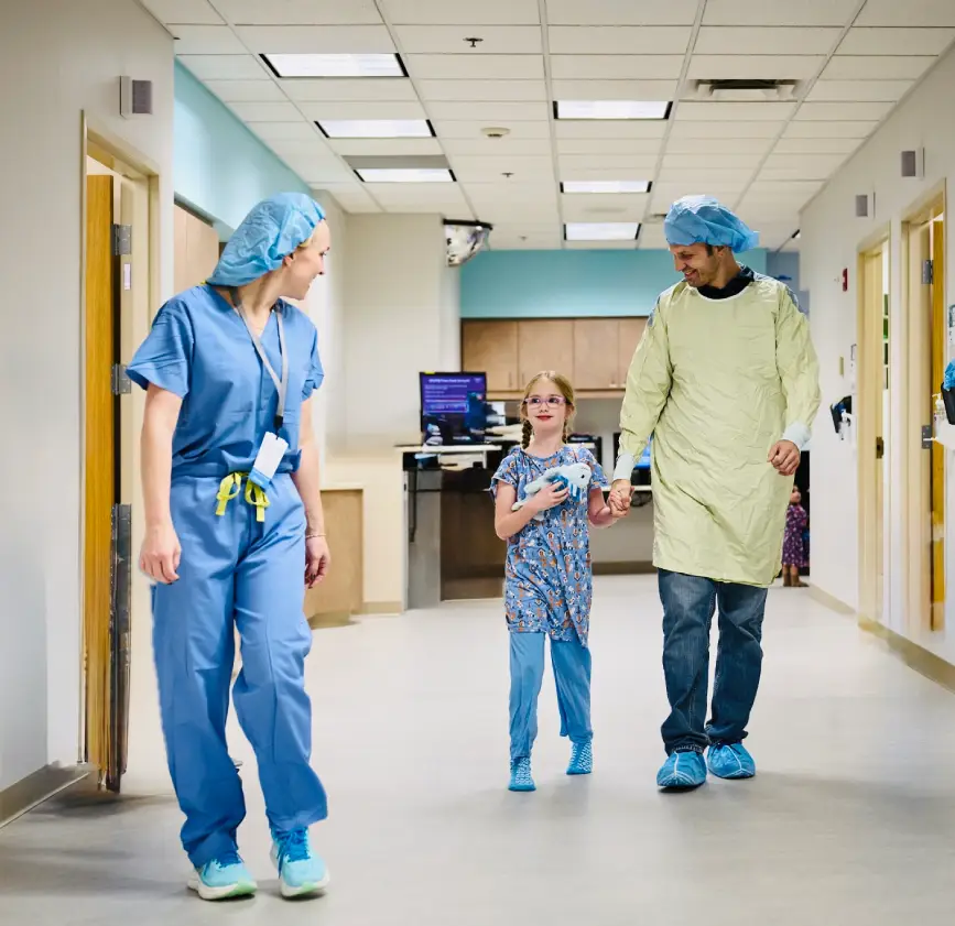 Child in hospital gown walking with parents and nurse in hallway.